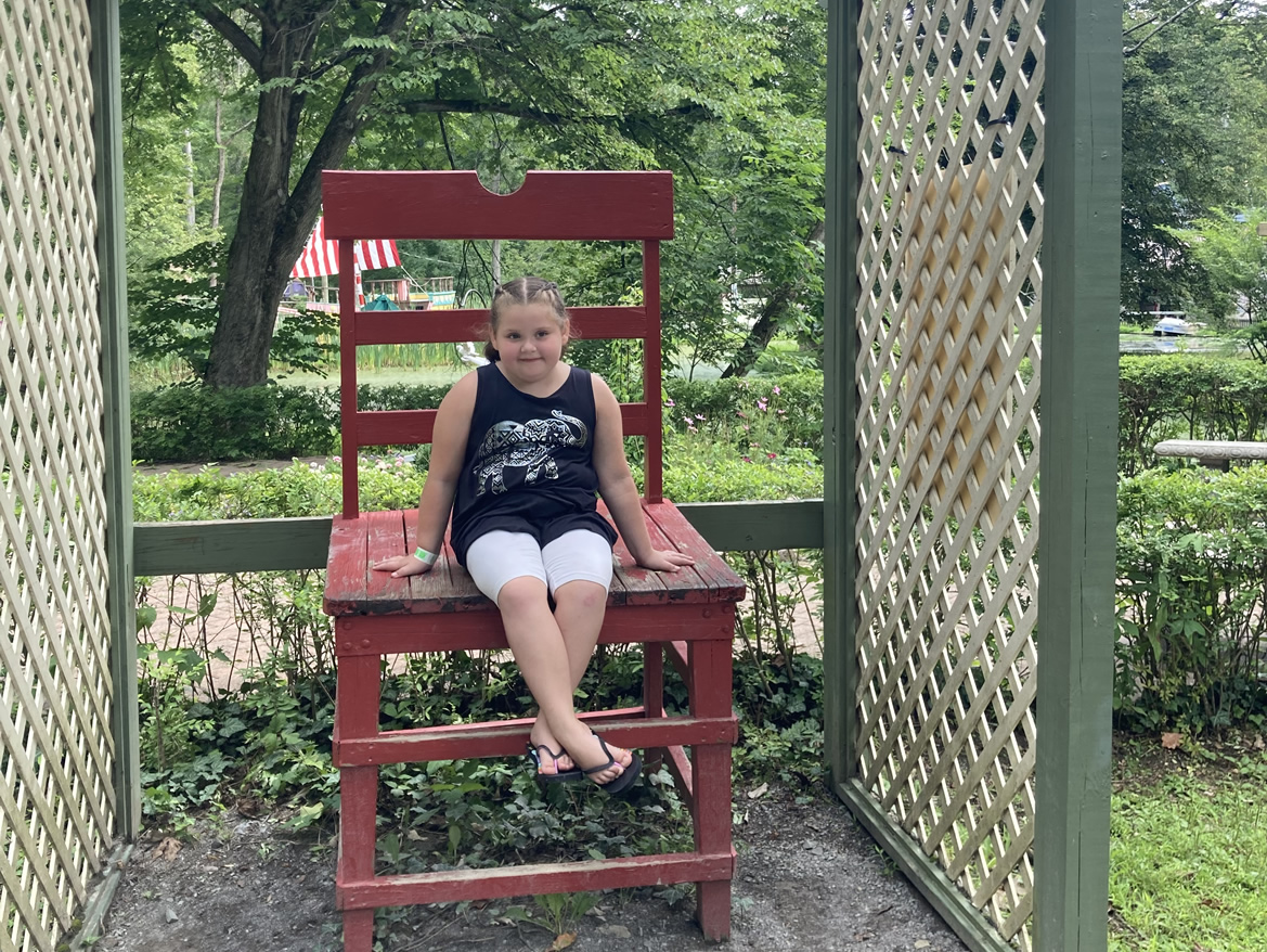 RaeLynn Cuppett sitting on an oversized wooden chair in front of a garden walkway under a large wooden terrance.